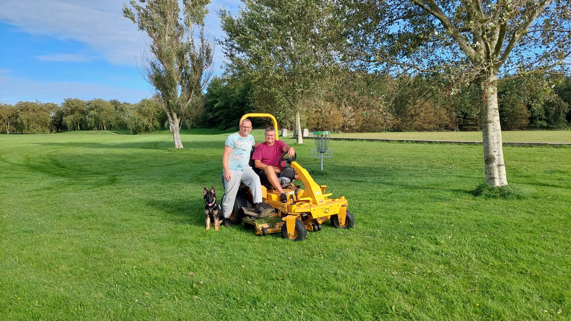 Jan en zijn collega op een grasmaaier op het grasveld waar discgolf wordt gespeeld met een hond naast de grasmaaier.